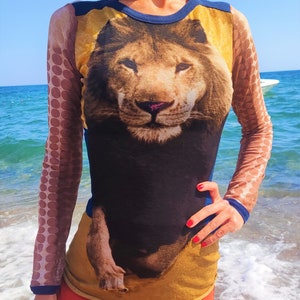 May include: A long-sleeved shirt with a lion graphic printed on the front. The shirt is made of a sheer, mesh fabric with brown polka dots on the sleeves. The lion is brown and black with a yellow background.