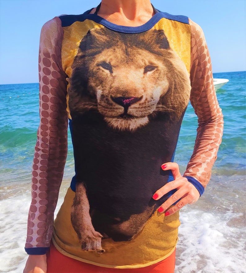 May include: A long-sleeved shirt with a lion graphic printed on the front. The shirt is made of a sheer, mesh fabric with brown polka dots on the sleeves. The lion is brown and black with a yellow background.