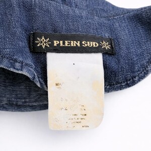 May include: Close-up of a denim garment with a black label featuring the text "PLEIN SUD" in gold lettering. The label is adorned with starburst designs on each side. A white tag with faded text is attached.