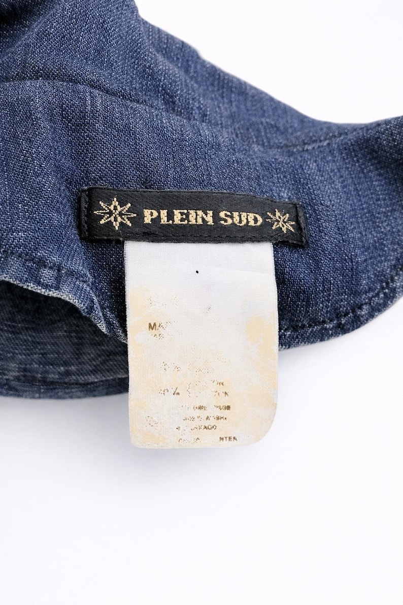 May include: Close-up of a denim garment with a black label featuring the text "PLEIN SUD" in gold lettering. The label is adorned with starburst designs on each side. A white tag with faded text is attached.