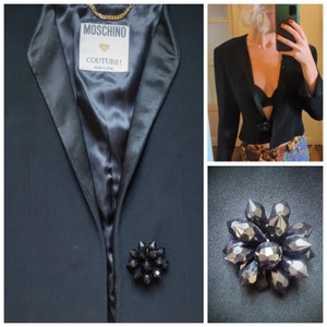 May include: A black blazer with a satin lapel and a black crystal brooch. The blazer has a label that says "Moschino Couture! Made in Italy."