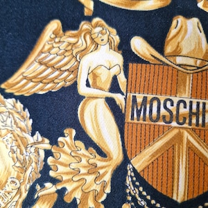 May include: A black fabric with a gold and orange design featuring a winged angel, a cowboy hat, and the word "MOSCHINO".