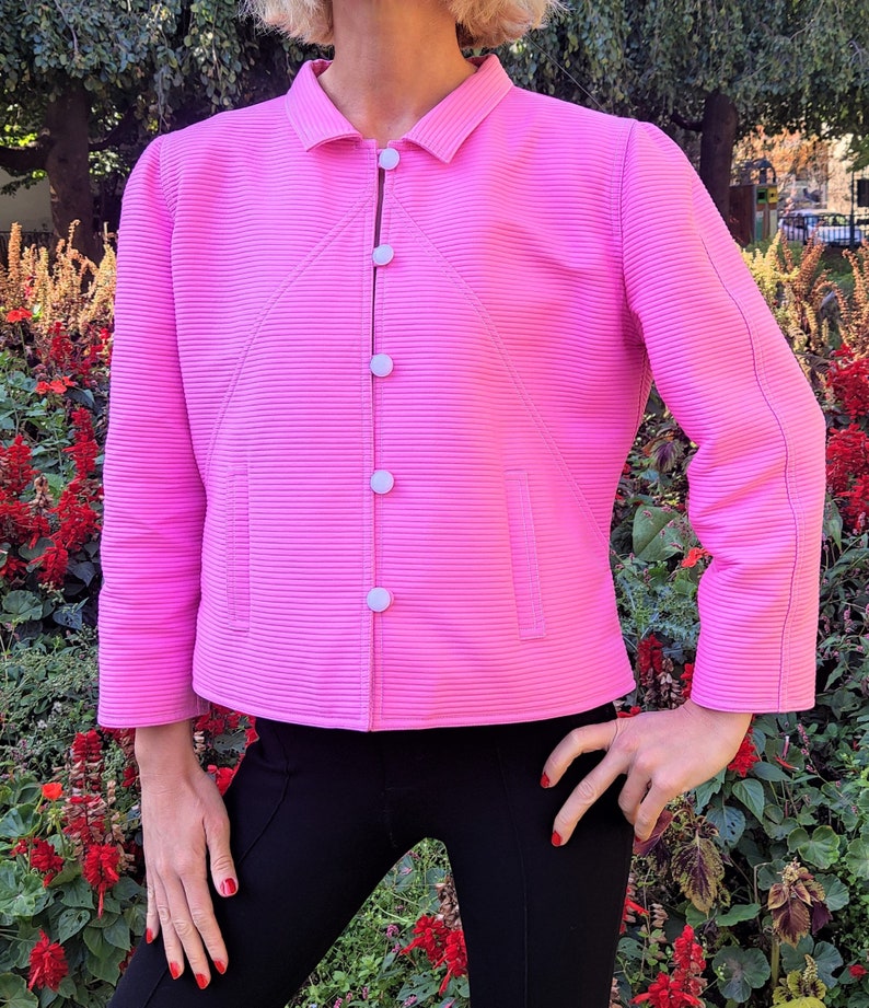 May include: A pink, ribbed, collared jacket with white buttons. The jacket has a slightly cropped length and a fitted silhouette.