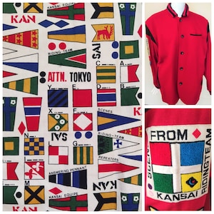 Może przedstawiać: Czerwono-czarna kurtka bejsbolowa z białym tłem, ozdobiona wzorem kolorowych flag z tekstem "KANSAI EQUIPE", "RIDING-TEAM", "ATTN. TOKYO", "SCENES", "REPEATERS", "ANSWERING PENNANT", "IVS", "FROM" i "KANSAI".