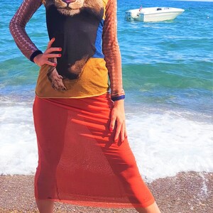 May include: A woman wearing a long-sleeved shirt with a lion print and a sheer orange skirt. She is standing on a beach with the ocean in the background.
