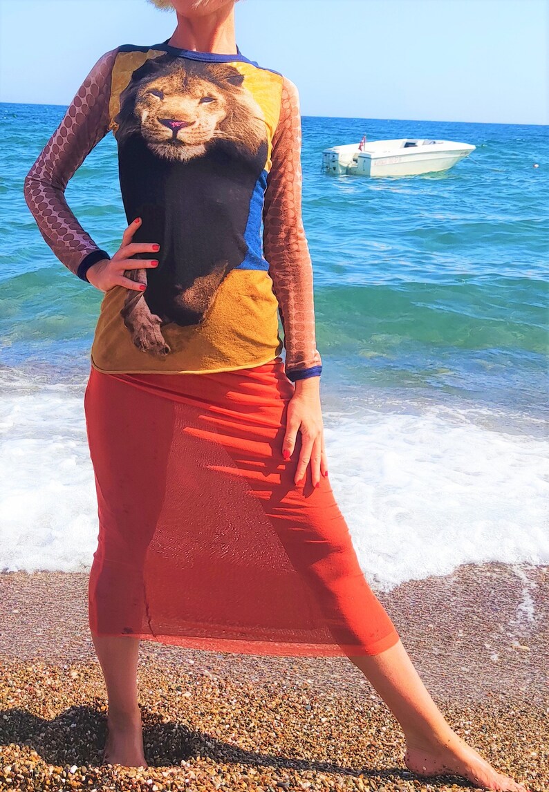 May include: A woman wearing a long-sleeved shirt with a lion print and a sheer orange skirt. She is standing on a beach with the ocean in the background.