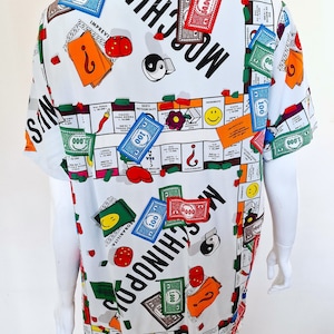 May include: A white short-sleeved shirt with a colorful Monopoly board game print. The shirt features the text "Moschino" and "Moschinop" repeated throughout the design.