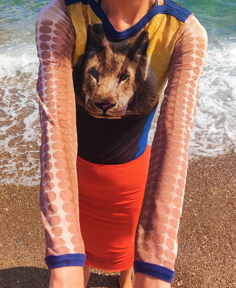 May include: A long-sleeved shirt with a lion print and polka dot sleeves. The shirt is yellow, blue, and brown. The sleeves are sheer and have a brown polka dot pattern. The shirt is paired with a red skirt.