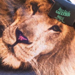 May include: Close-up of a lion's face printed on a black t-shirt. The t-shirt has a Jean Paul Gaultier Maille label attached to the collar. Made in Italy.