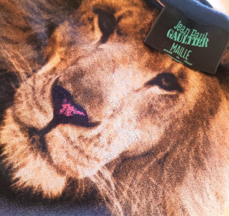 May include: Close-up of a lion's face printed on a black t-shirt. The t-shirt has a Jean Paul Gaultier Maille label attached to the collar. Made in Italy.