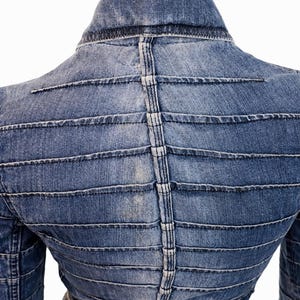 Può includere: Giacca di jeans blu chiaro con dettagli di cucitura orizzontali e verticali. La parte posteriore della giacca presenta un design unico con linee parallele e una cucitura verticale centrale. La giacca è esposta su un manichino.