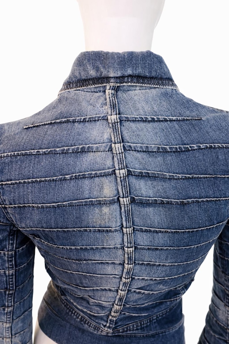 May include: A light blue denim jacket with horizontal and vertical stitched detailing. The back of the jacket features a unique design with parallel lines and a central vertical seam. The jacket is displayed on a mannequin.