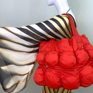 May include: A bright red puffer tote bag with a distinctive, rounded shape. The bag has a zipped pocket and silver-coloured hardware. A black and white patterned scarf is draped over the mannequin's arm, providing a contrasting element to the outfit.
