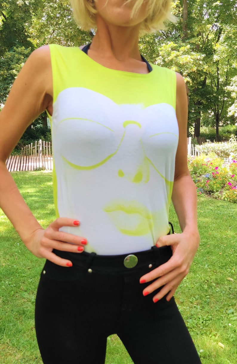 May include: A woman wearing a sleeveless shirt with a white graphic of a face wearing sunglasses. The shirt is neon yellow. The woman is wearing black pants.