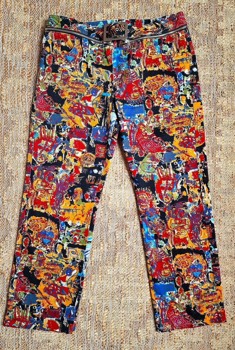 May include: Black pants with a colorful abstract print featuring a variety of shapes, lines, and figures. The pants have a zipper and a belt with a silver buckle.