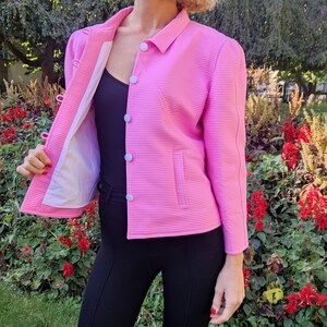 May include: A pink, ribbed, cropped jacket with white buttons and a single pocket. The jacket is unbuttoned and the lining is visible.