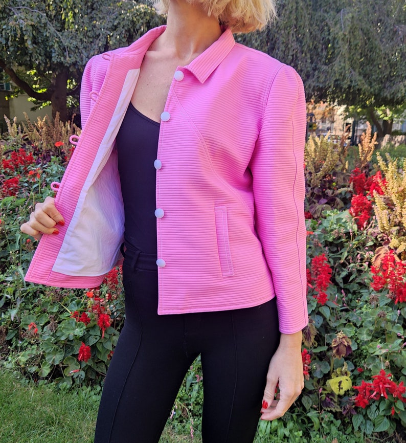 May include: A pink, ribbed, cropped jacket with white buttons and a single pocket. The jacket is unbuttoned and the lining is visible.