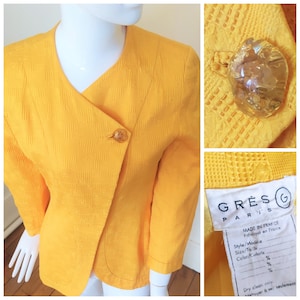 May include: A yellow blazer with a diagonal closure and a single button. The blazer has a textured fabric and a label that reads "GRES G PARIS MADE IN FRANCE Fabriqué en France Style/Modèle Size/Toile Color/Coloris % % % Dry clean only. nettoyer à sec seulement."