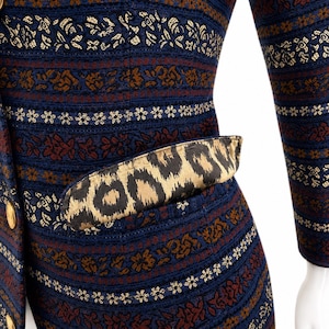 Puede incluir: Una chaqueta de manga larga con base azul oscuro y bandas horizontales de complejos patrones florales en oro y rojo intenso. La prenda incluye un bolsillo con una solapa de estampado de leopardo y botones redondos de metal dorado.
