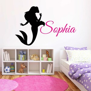 May include: Black silhouette of a mermaid with the name Sophia written in pink cursive lettering on a white wall. The wall is decorated with a white shelf with books, toys, and a phone.