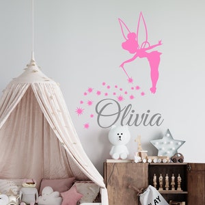 Puede incluir: Pegatina de pared de silueta de hada rosa con una varita y estrellas brillantes, con el nombre Olivia escrito debajo. La pegatina está en una pared blanca sobre una cama con dosel con un juego de cama blanco y rosa.