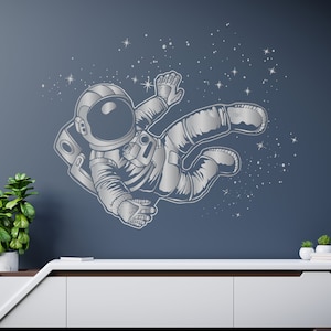 May include: A silver astronaut decal floating in space with a starry background. The astronaut is wearing a spacesuit and helmet. The decal is on a gray wall above a white dresser.
