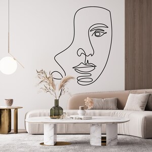 May include: A minimalist living room featuring a large black line art drawing of a face on a white wall. The room includes a beige sofa, a marble coffee table, and a gold side table, creating a modern and artistic ambiance.