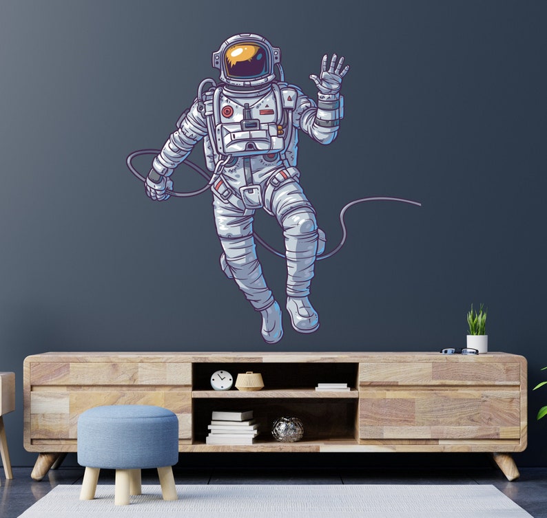 May include: A white astronaut with a blue and orange spacesuit is floating in space with a tether attached to their wrist. The astronaut is waving with their right hand. The background is a dark blue.