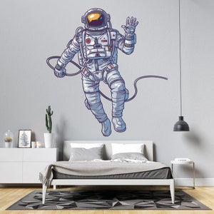 May include: A white astronaut with a blue and yellow helmet is floating in space with a cord attached to his back. He is waving with his right hand. The astronaut is on a light gray wall with a white bed in the foreground.