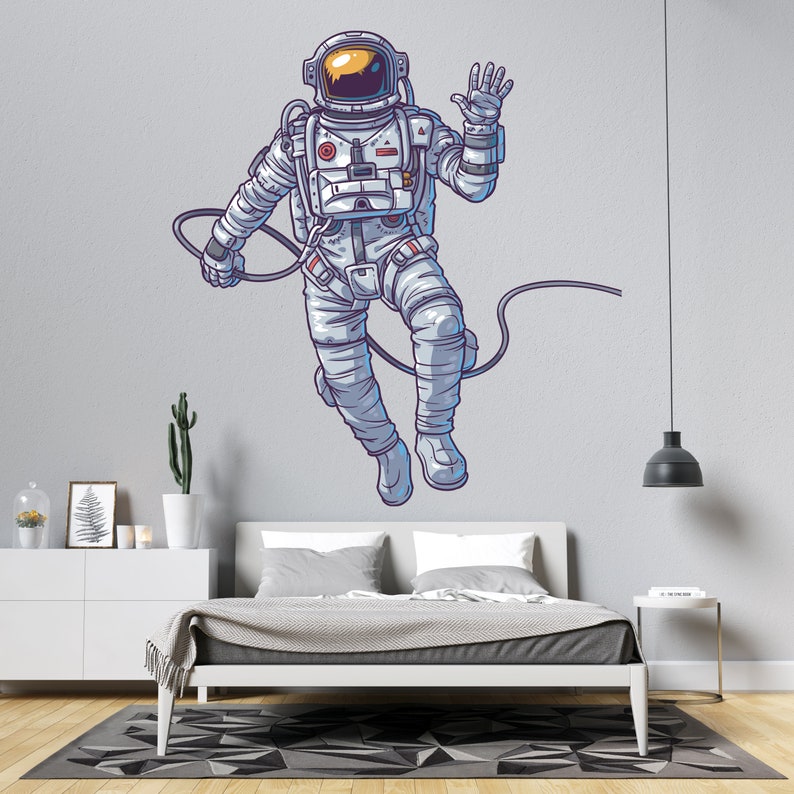 May include: A white astronaut with a blue and yellow helmet is floating in space with a cord attached to his back. He is waving with his right hand. The astronaut is on a light gray wall with a white bed in the foreground.