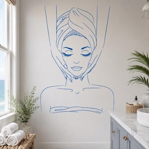 May include: A blue line art wall decal featuring a person with eyes closed, wrapped in a towel, receiving a facial massage. The design shows the person's head and shoulders with hands positioned on the sides of the face, rendered in simple strokes.