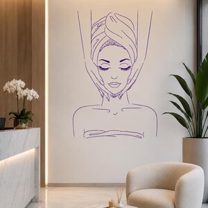 May include: A purple line art wall decal featuring a person with eyes closed, wrapped in a towel, receiving a facial massage from two hands. The minimalist graphic design depicts a serene spa treatment scene with clean, continuous outlines.