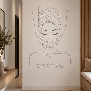 May include: A black line art wall decal featuring a person with eyes closed, wrapped in a towel, receiving a facial massage. The minimalist illustration depicts the head, shoulders, and hands of a practitioner performing the massage treatment.