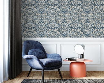 May include: A blue velvet armchair with black legs sits on a wood floor in front of a wall with a blue and white damask wallpaper pattern. A small round side table with a coral-colored top sits next to the chair. A black and white book and a silver lamp are on the table.