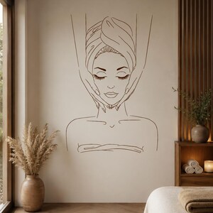 May include: A dark brown line art wall decal featuring a person with eyes closed, wrapped in a towel, receiving a facial massage from two hands. The minimalist illustration is centred on a light beige wall above a spa treatment room setting.