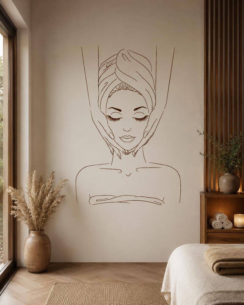 May include: A dark brown line art wall decal featuring a person with eyes closed, wrapped in a towel, receiving a facial massage from two hands. The minimalist illustration is centred on a light beige wall above a spa treatment room setting.