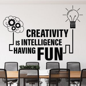 May include: Black and white wall decal with the text "Creativity is intelligence having fun". The decal features a lightbulb and a brain with gears. The decal is on a white wall in a conference room with chairs around a table.