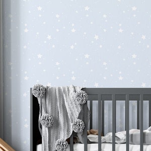 May include: A light blue baby crib with a white star pattern wallpaper. A gray knitted blanket with pom-poms is draped over the crib.