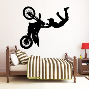 May include: Black silhouette wall decal of a motocross rider performing a stunt, with the motorcycle upside down. The decal is placed above a bed with a striped comforter and pillows. A red nightstand lamp is visible.