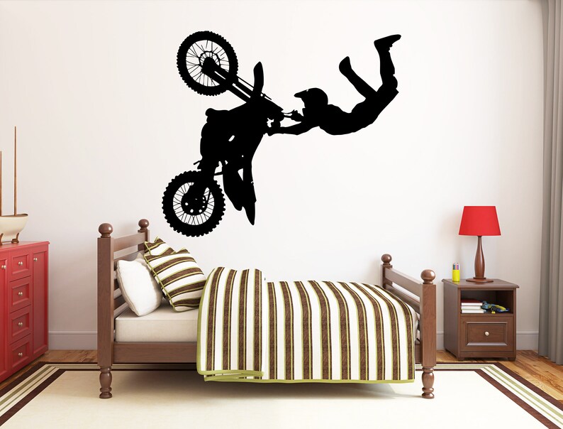 May include: Black silhouette wall decal of a motocross rider performing a stunt, with the motorcycle upside down. The decal is placed above a bed with a striped comforter and pillows. A red nightstand lamp is visible.