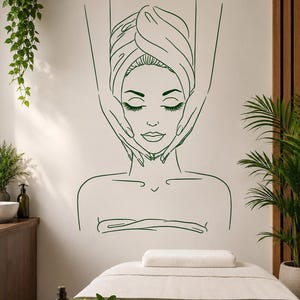 May include: A dark green line art wall decal depicting a person with eyes closed, wearing a towel wrapped around their head, while two hands gently massage the sides of their face. The minimalist graphic design features clean, continuous strokes.