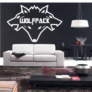 Pode incluir: Um logotipo branco de matilha de lobos com três cabeças de lobo em uma parede preta. O logotipo está acima de um sofá branco e uma mesa de café preta com um vaso e livros em cima.