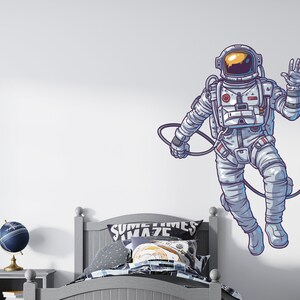 May include: A white wall with a cartoon illustration of an astronaut floating in space. The astronaut is wearing a white spacesuit with a blue visor and is waving. The astronaut is connected to a long, thin, black cord. The wall is also decorated with a bed with a gray headboard and a blue and white patterned pillow. The bed is in front of a nightstand with a blue globe on top.