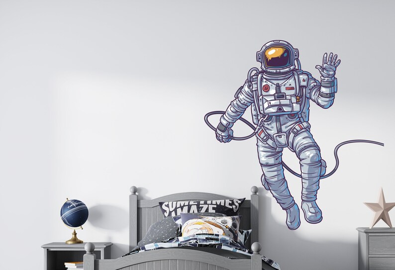 May include: A white wall with a cartoon illustration of an astronaut floating in space. The astronaut is wearing a white spacesuit with a blue visor and is waving. The astronaut is connected to a long, thin, black cord. The wall is also decorated with a bed with a gray headboard and a blue and white patterned pillow. The bed is in front of a nightstand with a blue globe on top.