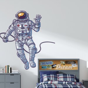 May include: A white astronaut wall decal with a blue outline, waving with a cord attached to its back. The decal is on a white wall above a gray headboard with a corkboard with the word "Dream" on it.