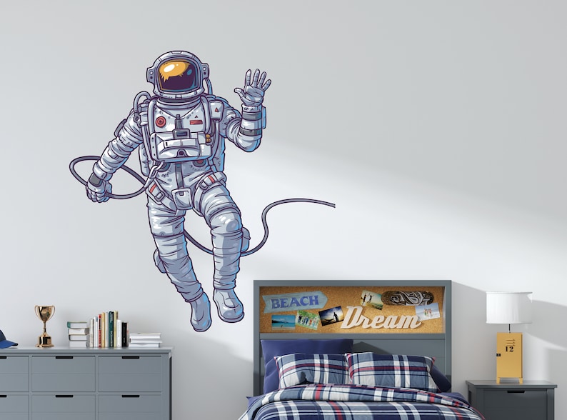 May include: A white astronaut wall decal with a blue outline, waving with a cord attached to its back. The decal is on a white wall above a gray headboard with a corkboard with the word "Dream" on it.