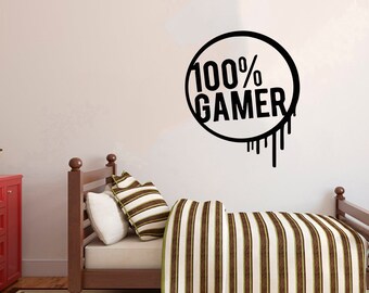 May include: A black and white wall decal with the text "100% GAMER" in a circle with drips coming from the bottom. The decal is on a white wall above a brown wooden bed with a striped bedspread.