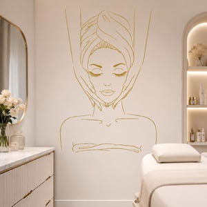 May include: A gold line art wall decal featuring a stylised illustration of a person receiving a facial massage. The design depicts a face with closed eyes, a towel wrapped around the head, and hands positioned on the cheeks and jawline.