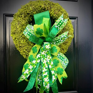 May include: A round moss wreath with a large bow featuring green ribbons with shamrock and floral patterns. The bow has a glittery green ribbon and is centered on the wreath, creating a festive St. Patrick's Day decoration.