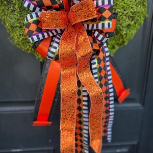 May include: A decorative bow with orange, black, and white ribbons. The bow features a mix of patterns, including harlequin diamonds and stripes. The center ribbon is a shimmering orange, and the bow is attached to a green wreath, perfect for Halloween.